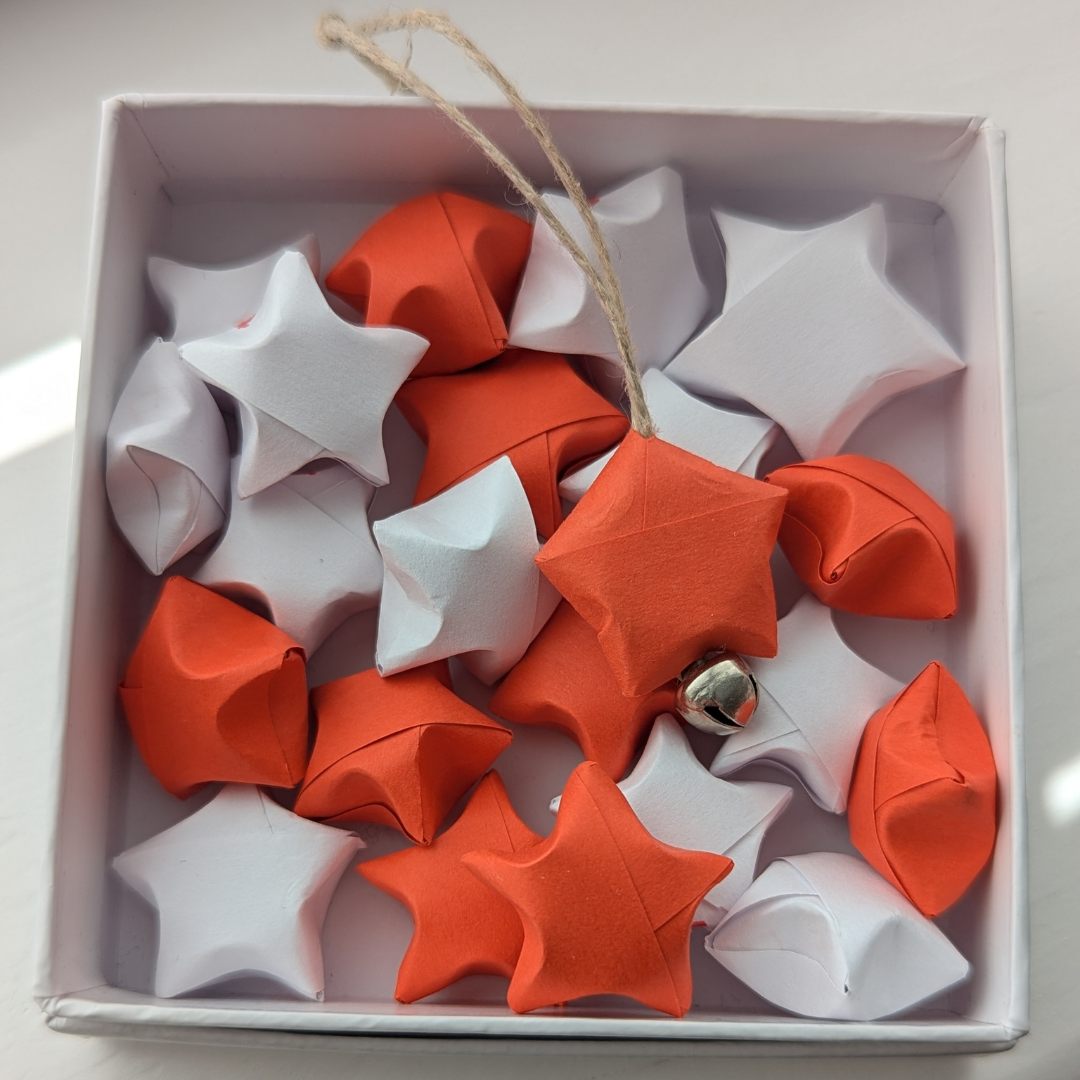 Image/png close up of a small white box, containing a mix of red and white paper 'puffed' stars origami style. One has a small bell and string attached.