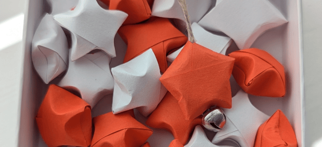 Image/png close up of a small white box, containing a mix of red and white paper 'puffed' stars origami style. One has a small bell and string attached.